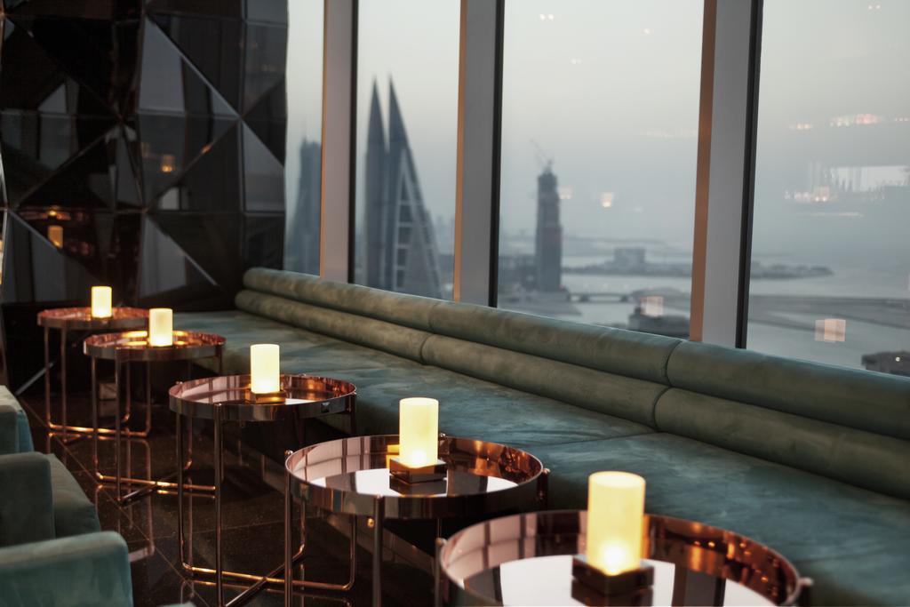 מנאמה The Domain Bahrain Hotel And Spa - Adults Friendly 16 Years Plus מראה חיצוני תמונה The photo depicts a stylish lounge area with a modern aesthetic. There are several circular tables made of metallic materials, each topped with a small glowing candle. A long, plush seating area in a teal color runs along the window, offering a view 